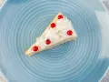 Kawałek pysznego ciasta z białą czekoladą, udekorowany bitą śmietaną i czerwonymi porzeczkami, na niebieskim talerzu.