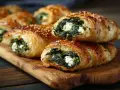 Chrupiące ciasto francuskie ze szpinakiem i serem feta, posypane sezamem, ułożone na drewnianej desce.