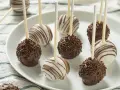 Słodkie cake pops na patyczkach, niektóre w czekoladzie z posypką, inne w białej polewie z czekoladowymi paskami.