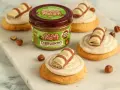 Pyszne ciasteczka inspirowane przepisem na crumbl cookies, z kremem i kawałkami czekolady. Słoik pasty z orzechów laskowych w tle.