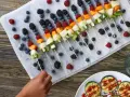Kolorowe szaszłyki owocowe, idealne przekąski dla dzieci. Na marmurowej desce ułożone borówki, maliny, jeżyny, melon, kiwi i morele.