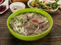 A miseczka aromatycznej zupy pho z wołowiną, makaronem ryżowym, świeżą kolendrą i chili. Obok sosy i dodatki.