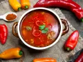 Aromaticzna zupa gulaszowa w misce, otoczona papryczkami chili i słodkimi paprykami. Na łyżce mielona papryka.
