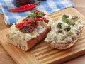 Dwa kawałki bagietki z twarogiem, suszonymi pomidorami i kaparami. Kapary dodają wyrazistego smaku.