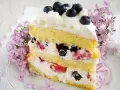 Pyszny tort z bitą śmietaną i mascarpone, przełożony owocami leśnymi, ozdobiony jagodami i kwitnącym bzem.