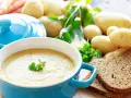 Aksamitna zupa ziemniaczana w niebieskiej misce, udekorowana natką pietruszki. Obok surowe ziemniaki, marchewki i kromki ciemnego chleba.