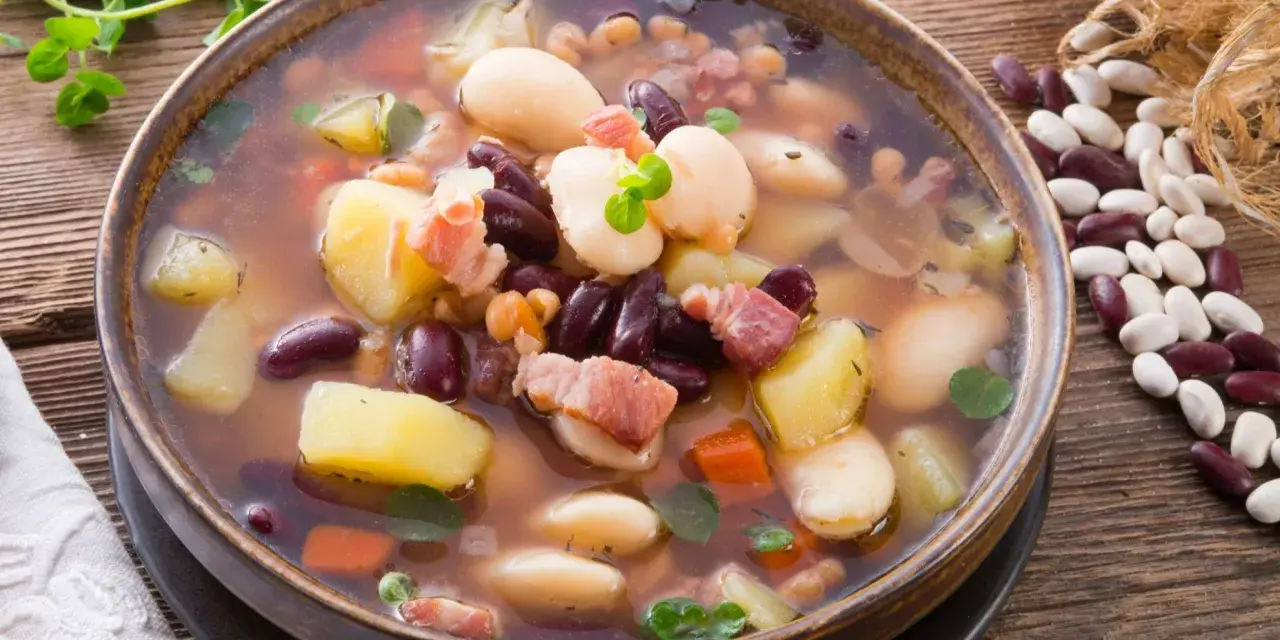 Gęsta, aromatyczna zupa fasolowa z ziemniakami i boczkiem, podana w glinianej misce. Obok leżą surowe fasole i gałązka ziół.