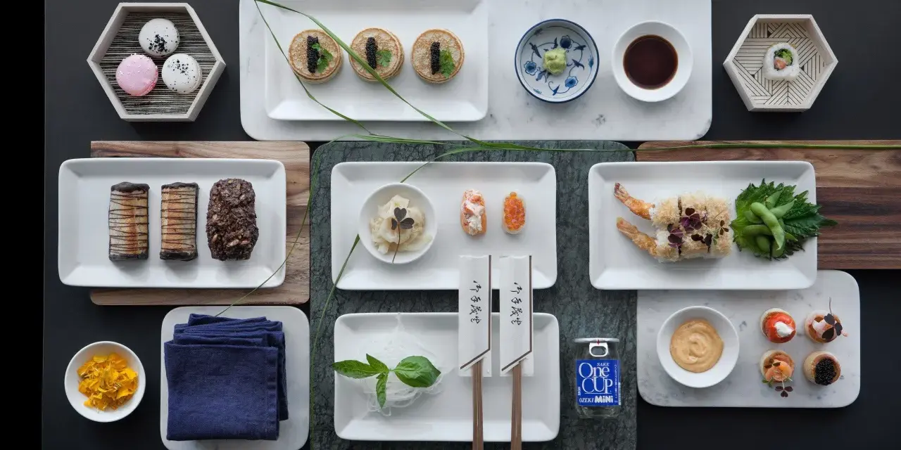 Elegancka aranżacja stołu z różnorodnymi przekąskami, w tym mini naleśnikami z kawiorem, sushi, krewetkami w tempurze i makaronikami. Idealne na wieczór, gdy masz ochotę na coś pysznego do sushi.