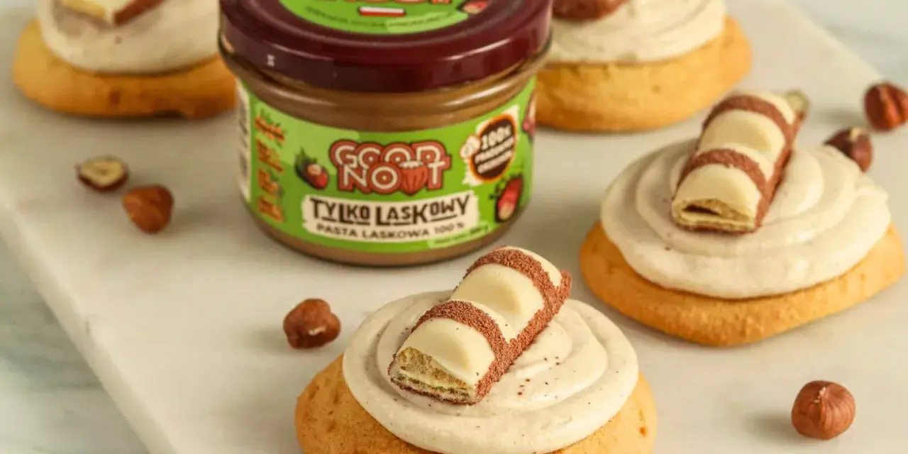 Pyszne ciasteczka inspirowane przepisem na crumbl cookies, z kremem i kawałkami czekolady. Słoik pasty z orzechów laskowych w tle.