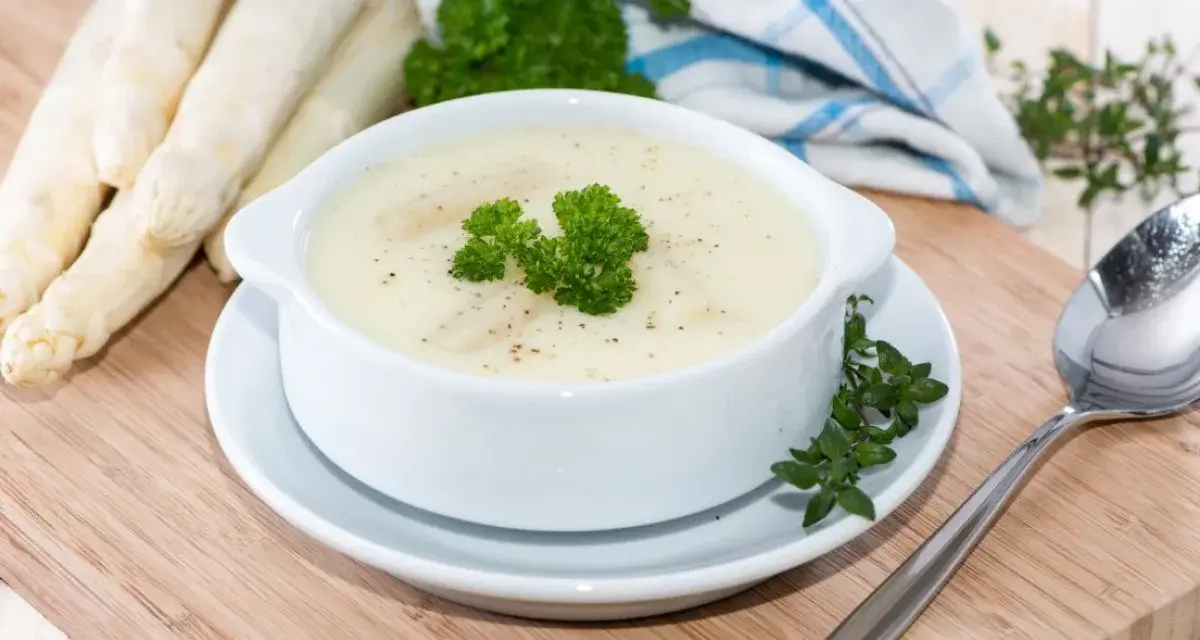 Aksamitna zupa szparagowa, udekorowana natką pietruszki, obok świeżych szparagów i łyżki.