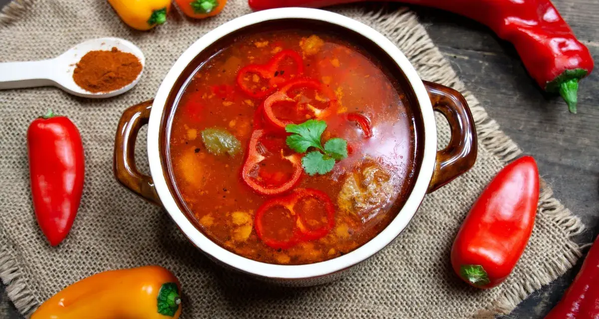 Aromaticzna zupa gulaszowa w misce, otoczona papryczkami chili i słodkimi paprykami. Na łyżce mielona papryka.