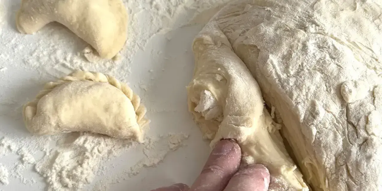Ręce formują ciasto na pierogi, tworząc półksiężycowate kształty. Mąka rozsypana na blacie.