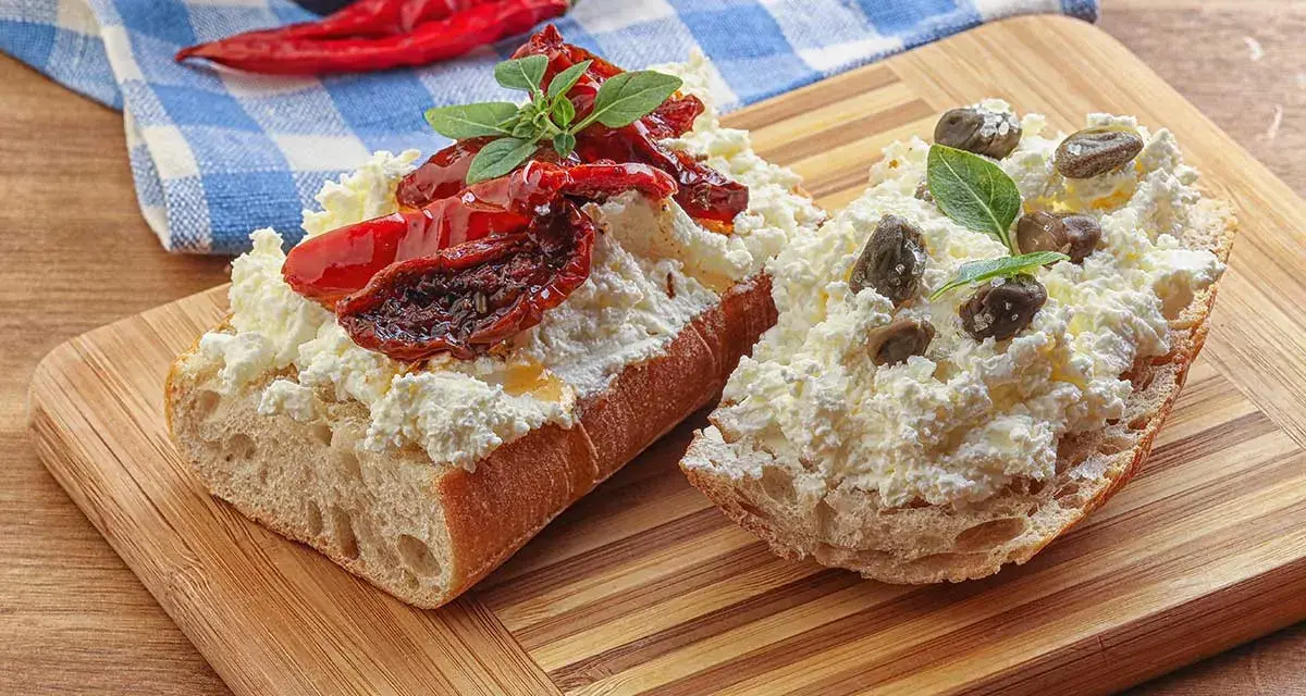 Dwa kawałki bagietki z twarogiem, suszonymi pomidorami i kaparami. Kapary dodają wyrazistego smaku.
