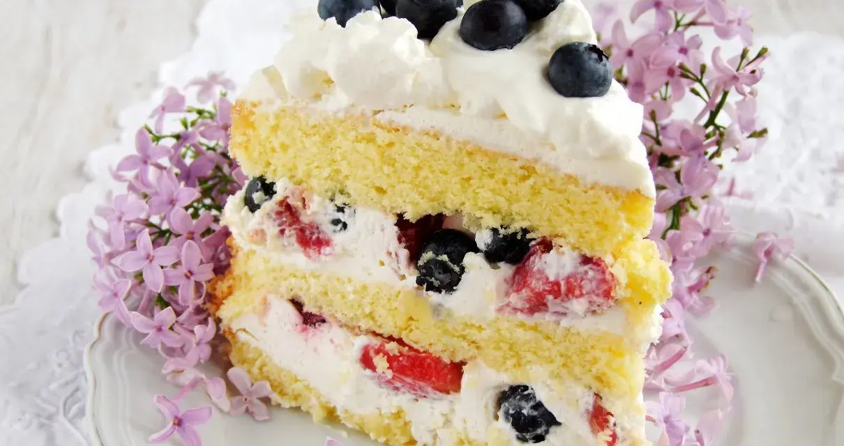Pyszny tort z bitą śmietaną i mascarpone, przełożony owocami leśnymi, ozdobiony jagodami i kwitnącym bzem.