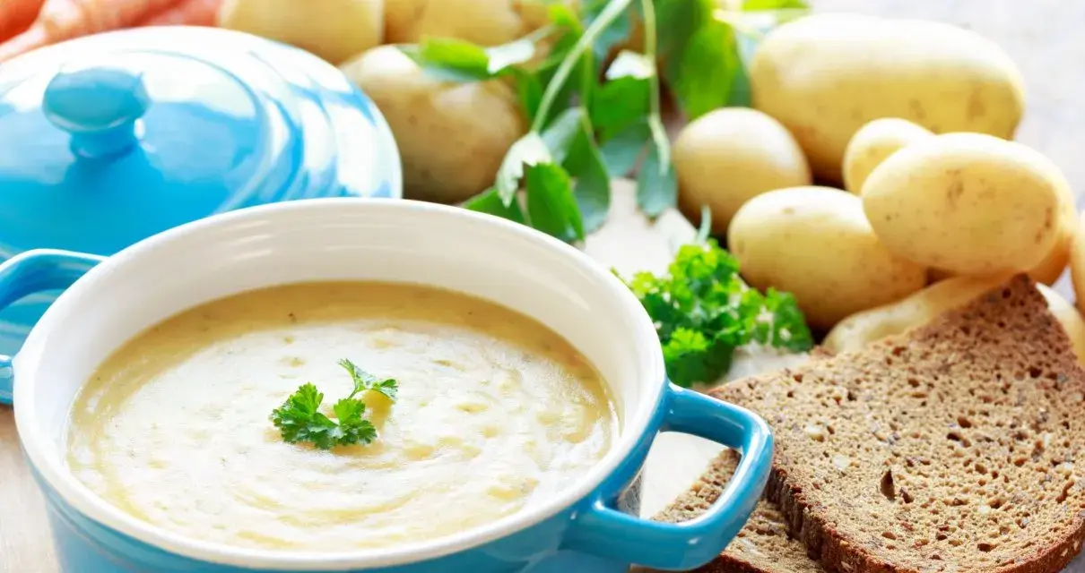 Aksamitna zupa ziemniaczana w niebieskiej misce, udekorowana natką pietruszki. Obok surowe ziemniaki, marchewki i kromki ciemnego chleba.