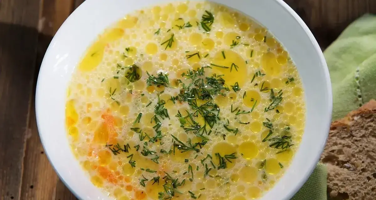 Aromaticzna zupa ogórkowa, pełna koperku i pływających kuleczek tłuszczu, czeka na talerzu obok kromki chleba.