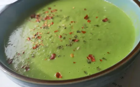 Aksamitna zupa z zielonego groszku, posypana płatkami chili i ziołami, w niebieskiej misce.