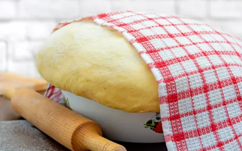 Puszyste ciasto drożdżowe rośnie w misce, przykryte kratkowaną ściereczką. Obok wałek do ciasta i rozsypana mąka.