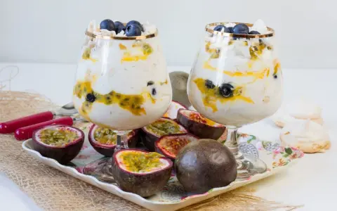 Dwa puchary z Eton Mess, przełożone kremem, marakują i borówkami, ozdobione bezą. Obok owoce marakui i bezy.