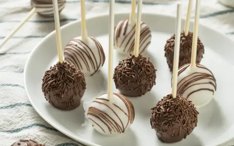 Słodkie cake pops na patyczkach, niektóre w czekoladzie z posypką, inne w białej polewie z czekoladowymi paskami.
