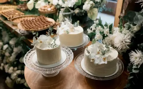 Elegancki, prosty tort na chrzest ozdobiony białymi kwiatami i zielenią. W tle słodki stół z deserami.