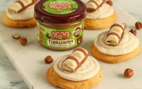 Pyszne ciasteczka inspirowane przepisem na crumbl cookies, z kremem i kawałkami czekolady. Słoik pasty z orzechów laskowych w tle.