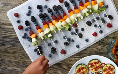 Kolorowe szaszłyki owocowe, idealne przekąski dla dzieci. Na marmurowej desce ułożone borówki, maliny, jeżyny, melon, kiwi i morele.