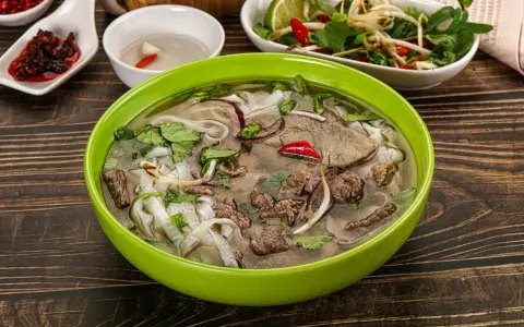 A miseczka aromatycznej zupy pho z wołowiną, makaronem ryżowym, świeżą kolendrą i chili. Obok sosy i dodatki.