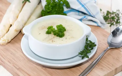 Aksamitna zupa szparagowa, udekorowana natką pietruszki, obok świeżych szparagów i łyżki.