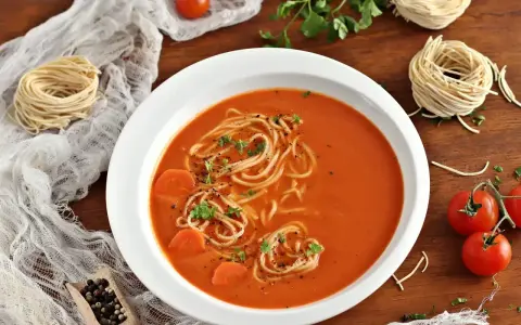 Aksamitna zupa pomidorowa z makaronem i marchewką, udekorowana natką pietruszki. Obok leżą surowe nitki makaronu i pomidorki.