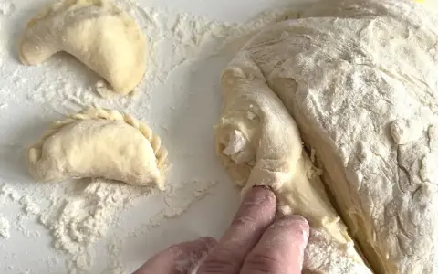 Ręce formują ciasto na pierogi, tworząc półksiężycowate kształty. Mąka rozsypana na blacie.