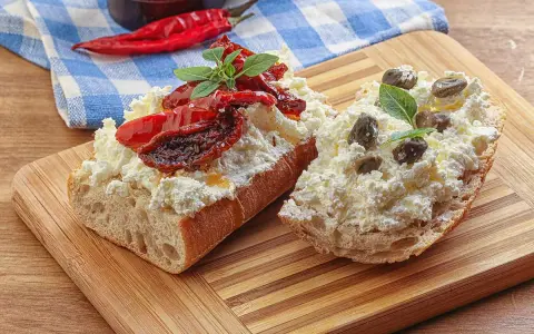 Dwa kawałki bagietki z twarogiem, suszonymi pomidorami i kaparami. Kapary dodają wyrazistego smaku.