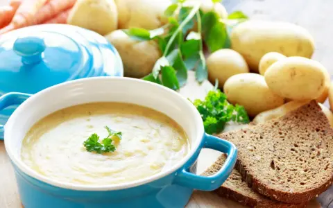 Aksamitna zupa ziemniaczana w niebieskiej misce, udekorowana natką pietruszki. Obok surowe ziemniaki, marchewki i kromki ciemnego chleba.