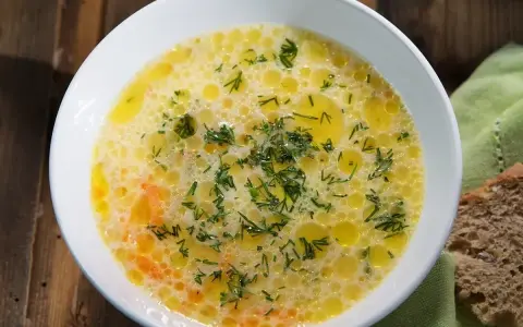 Aromaticzna zupa ogórkowa, pełna koperku i pływających kuleczek tłuszczu, czeka na talerzu obok kromki chleba.