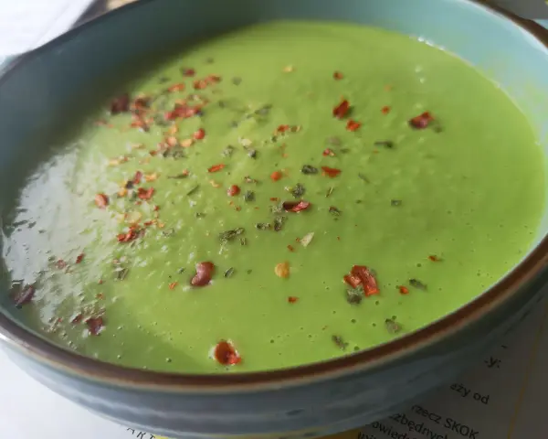 Aksamitna zupa z zielonego groszku, posypana płatkami chili i ziołami, w niebieskiej misce.