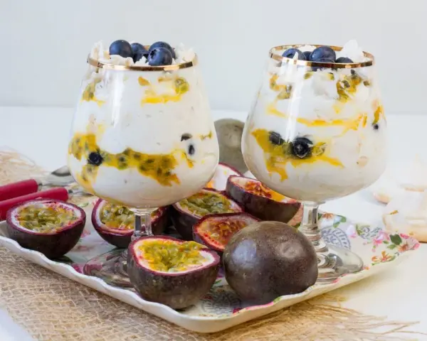 Dwa puchary z Eton Mess, przełożone kremem, marakują i borówkami, ozdobione bezą. Obok owoce marakui i bezy.