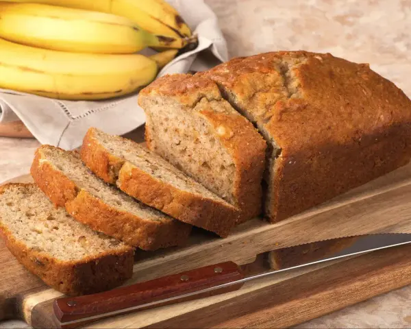 Pyszny, domowy chlebek bananowy, pokrojony na kromki, leży obok dojrzałych bananów.
