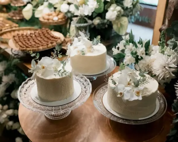 Elegancki, prosty tort na chrzest ozdobiony białymi kwiatami i zielenią. W tle słodki stół z deserami.