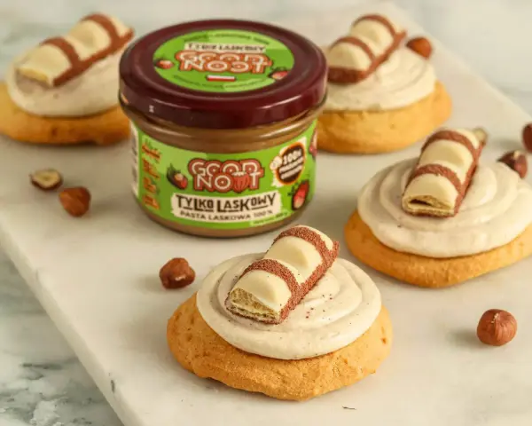 Pyszne ciasteczka inspirowane przepisem na crumbl cookies, z kremem i kawałkami czekolady. Słoik pasty z orzechów laskowych w tle.