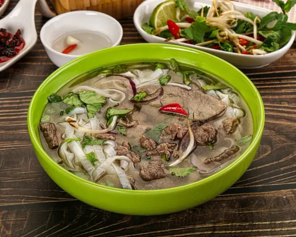 A miseczka aromatycznej zupy pho z wołowiną, makaronem ryżowym, świeżą kolendrą i chili. Obok sosy i dodatki.