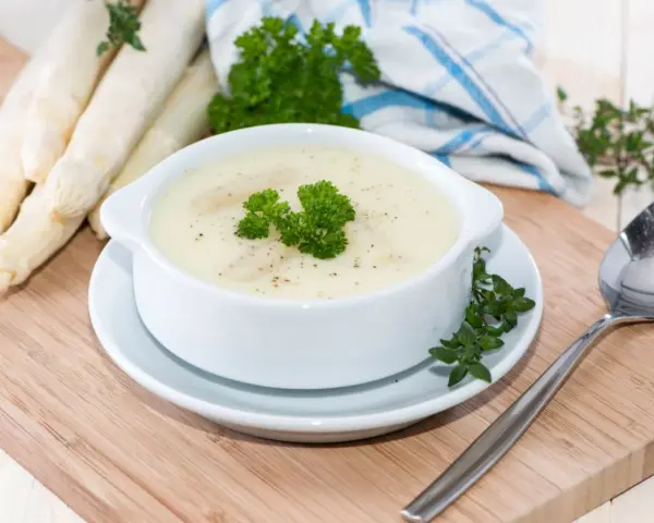 Aksamitna zupa szparagowa, udekorowana natką pietruszki, obok świeżych szparagów i łyżki.