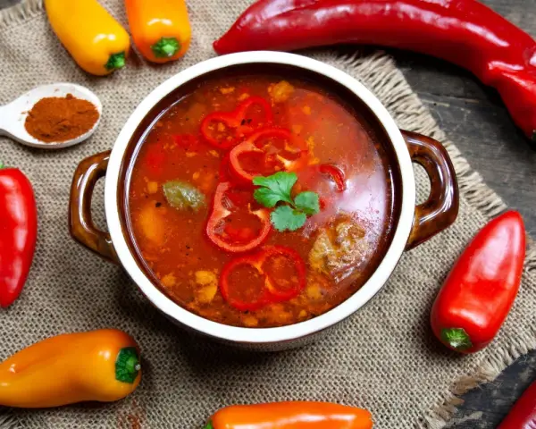 Aromaticzna zupa gulaszowa w misce, otoczona papryczkami chili i słodkimi paprykami. Na łyżce mielona papryka.