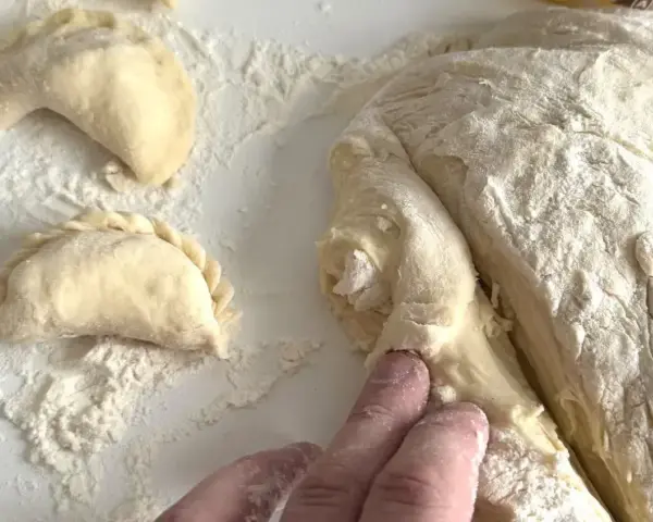 Ręce formują ciasto na pierogi, tworząc półksiężycowate kształty. Mąka rozsypana na blacie.