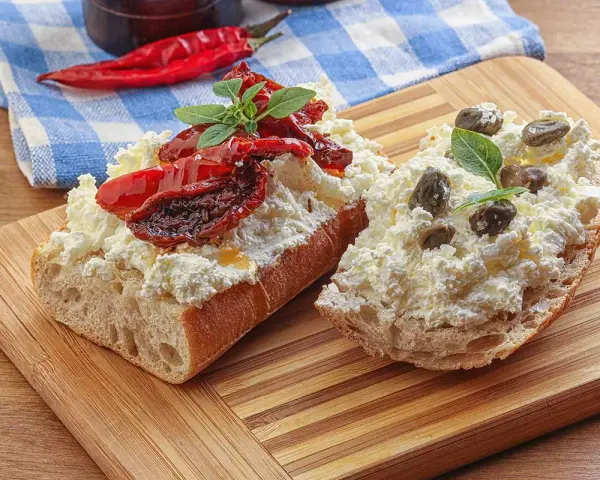 Dwa kawałki bagietki z twarogiem, suszonymi pomidorami i kaparami. Kapary dodają wyrazistego smaku.