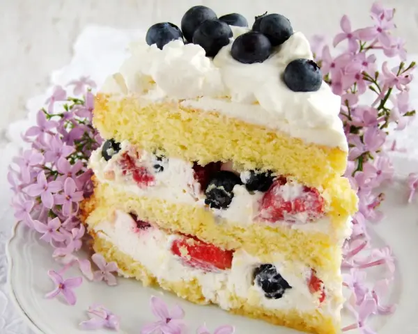 Pyszny tort z bitą śmietaną i mascarpone, przełożony owocami leśnymi, ozdobiony jagodami i kwitnącym bzem.