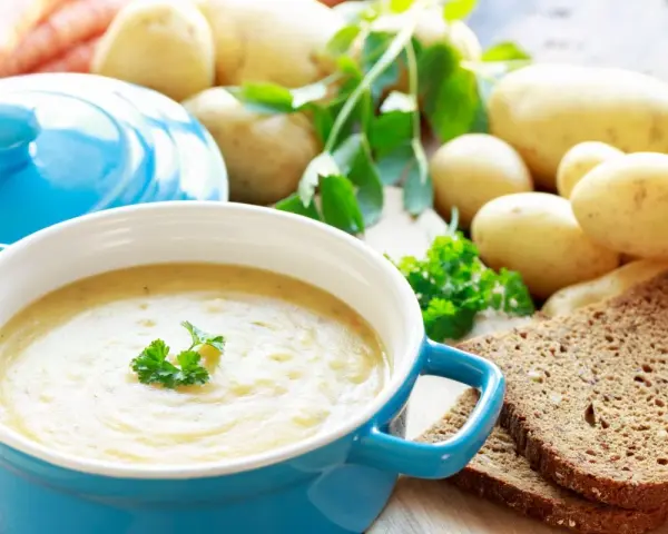 Aksamitna zupa ziemniaczana w niebieskiej misce, udekorowana natką pietruszki. Obok surowe ziemniaki, marchewki i kromki ciemnego chleba.