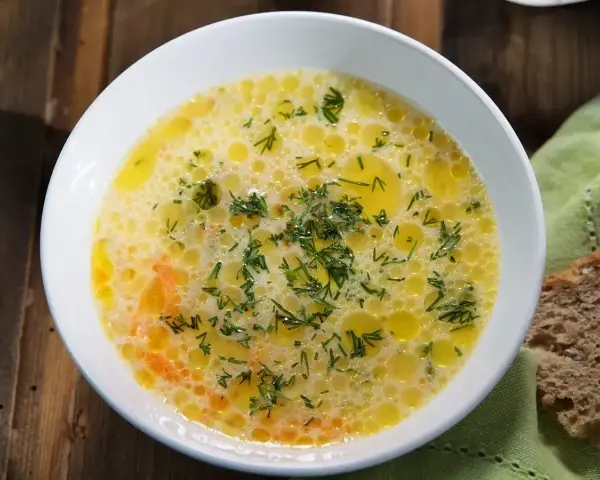 Aromaticzna zupa ogórkowa, pełna koperku i pływających kuleczek tłuszczu, czeka na talerzu obok kromki chleba.