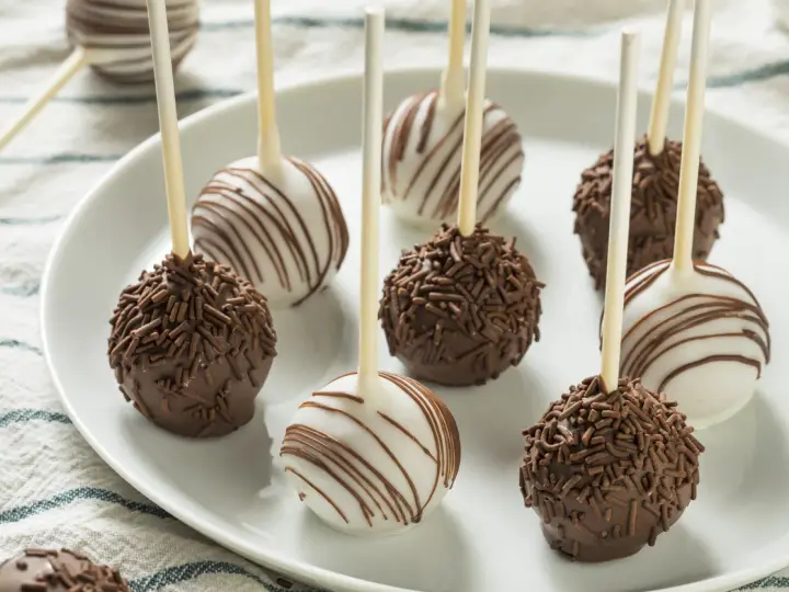 Słodkie cake pops na patyczkach, niektóre w czekoladzie z posypką, inne w białej polewie z czekoladowymi paskami.