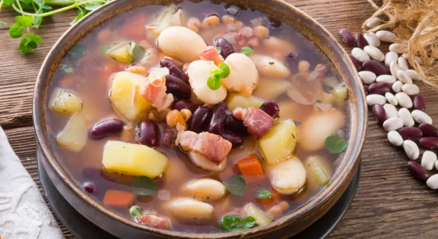 Gęsta, aromatyczna zupa fasolowa z ziemniakami i boczkiem, podana w glinianej misce. Obok leżą surowe fasole i gałązka ziół.