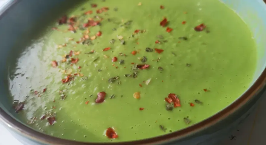 Aksamitna zupa z zielonego groszku, posypana płatkami chili i ziołami, w niebieskiej misce.